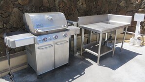 Ein Weber-Grill aus Edelstahl mit einem Beistelltisch vor einer Steinsäule.