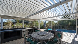 An outdoor patio with a wooden table and chairs, a grill, and a pool.