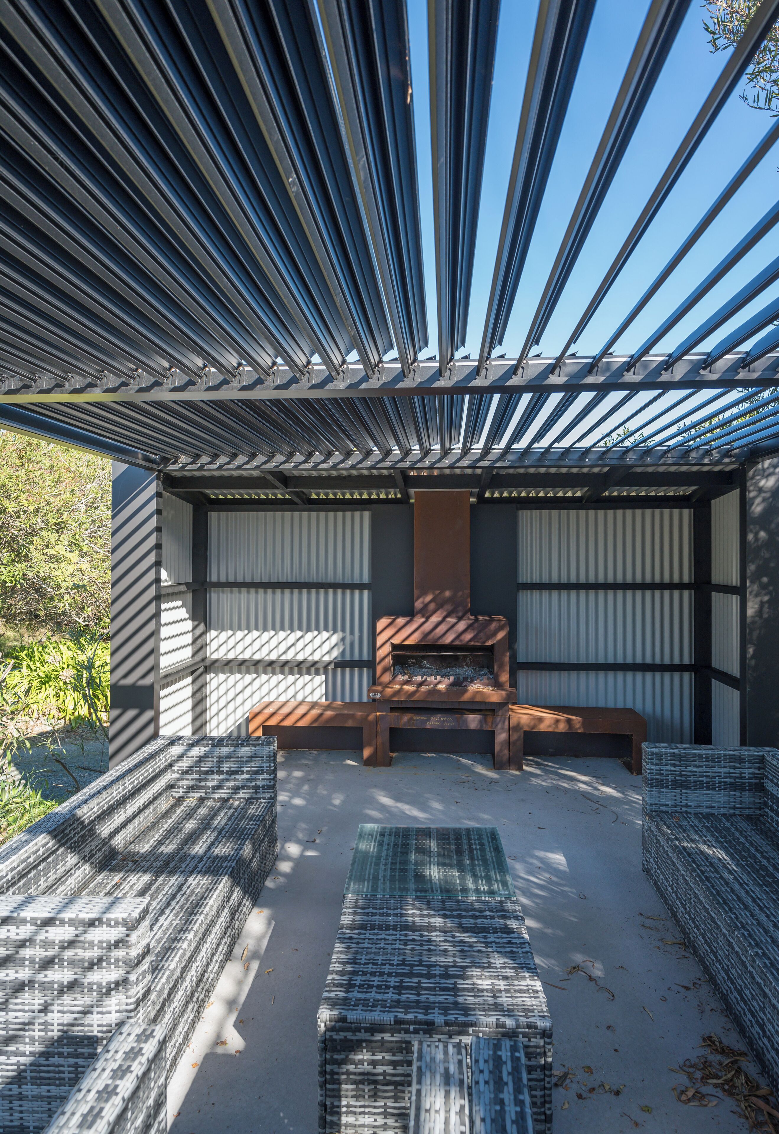A modern outdoor seating area with a bench and woven stools, a fire feature, and a metal pergola structure.