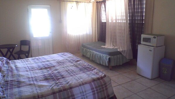 Una habitación pequeña y sencilla con cama, silla, mesa, microondas y refrigerador.
