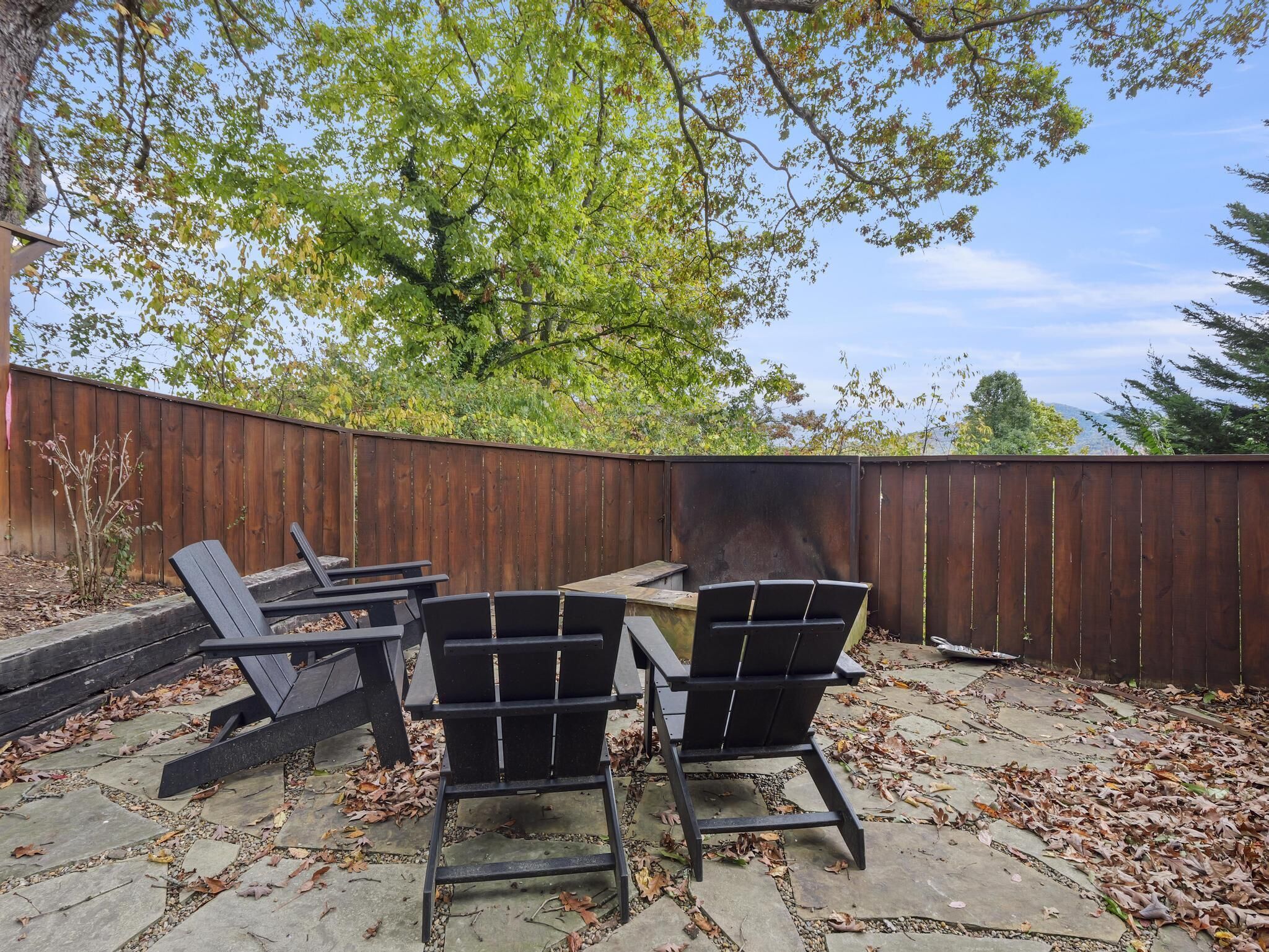Une terrasse extérieure avec une table en pierre et des chaises noires, entourée d’une clôture en bois et d’arbres.