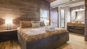 A bedroom with a large bed, wooden walls, a nightstand with a lamp, a vanity with a mirror, and a window providing natural light.