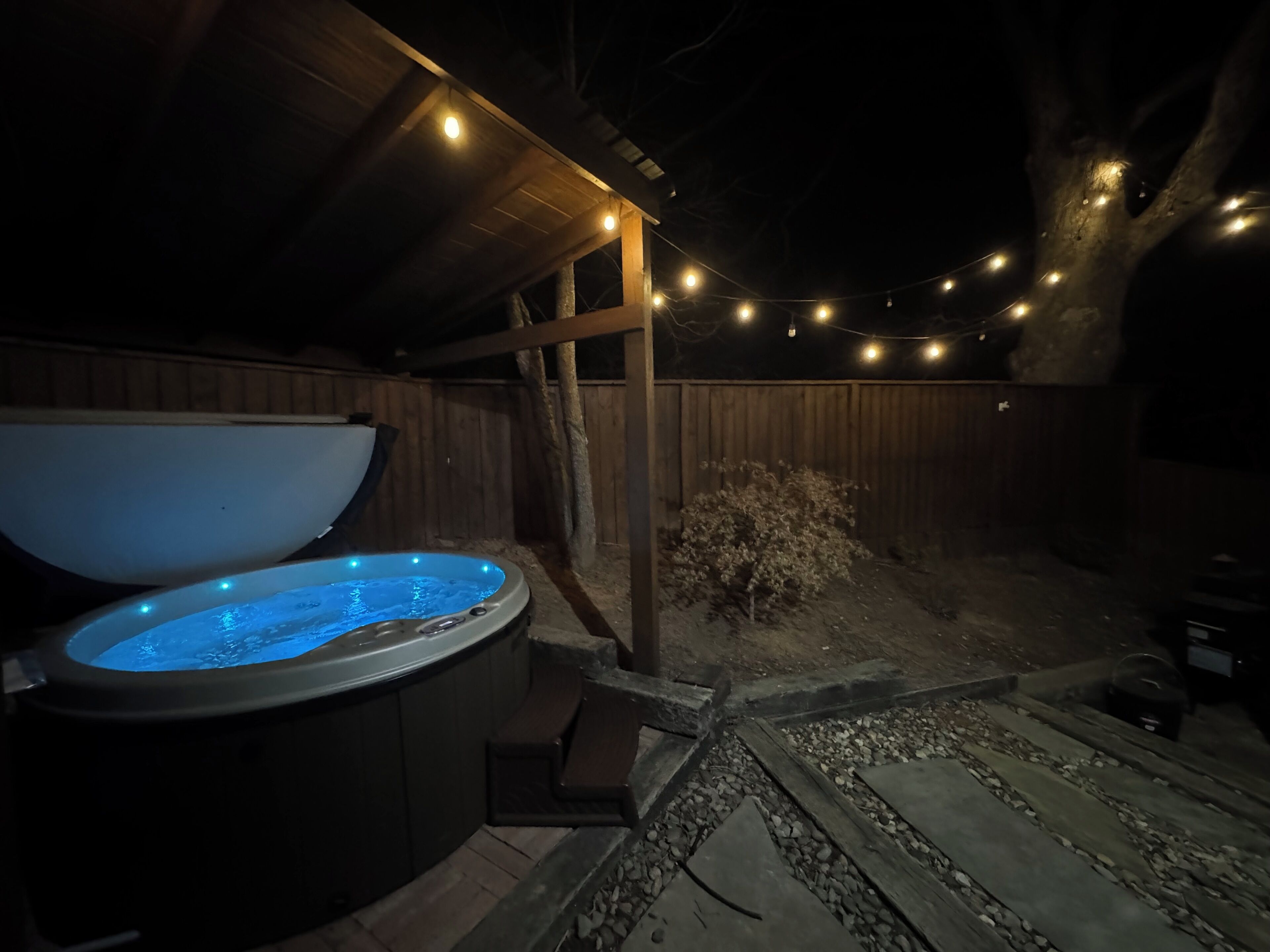 Ein Whirlpool unter einer Holzpergola mit Lichterketten, umgeben von einem Holzzaun und einem kleinen Busch.