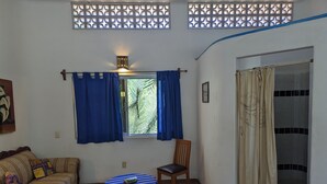 A room with a sofa, a small table, a chair, a window with blue curtains, and a view of greenery outside.