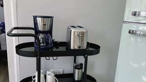 Een keukenhoek met een zwart metalen rek waarop een koffiezetapparaat, een broodrooster en een waterkoker staan. Een witte koelkast bevindt zich rechts.