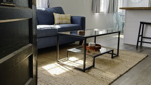 Een moderne woonkamer met een donkerhouten deur, een blauwe bank, een glazen salontafel en een keuken in de achtergrond.