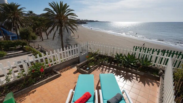 Vista sulla spiaggia con due lettini, una recinzione bianca e palme.