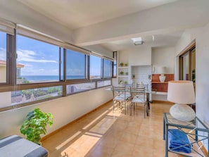 En balkon med udsigt over havet, et spiseområde med stole og et bord, samt et tekøkken med en lampe og hylder.