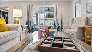 Una sala de estar moderna con una mesa de centro de vidrio, un sofá, un sillón y una lámpara. Se ve el exterior a través de un ventanal.