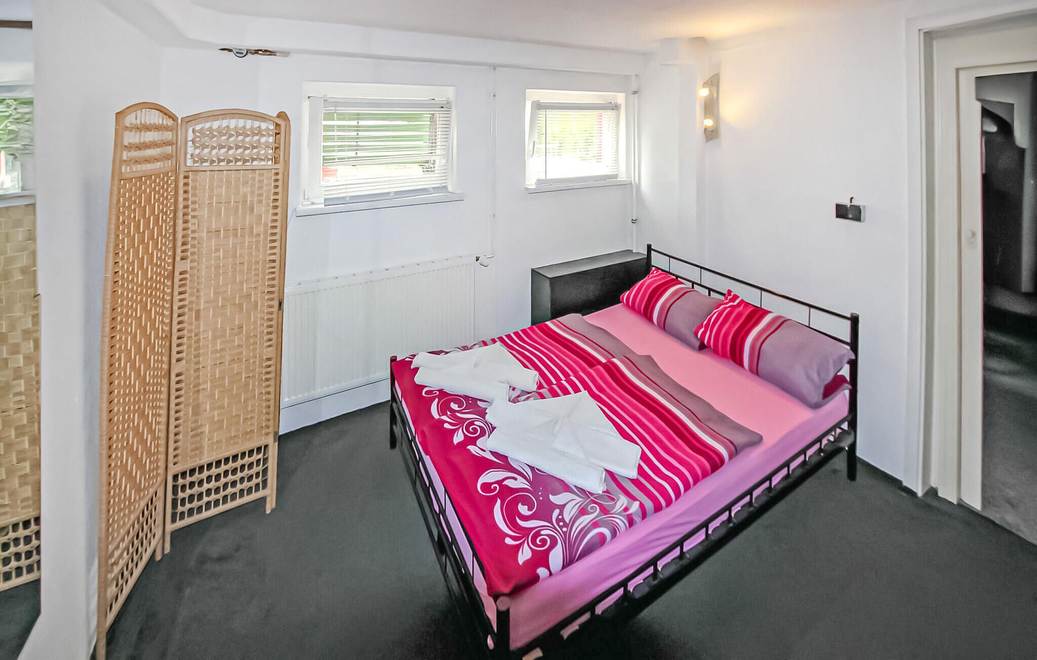 A bedroom with a bed, two windows with blinds, a radiator, and a wardrobe.