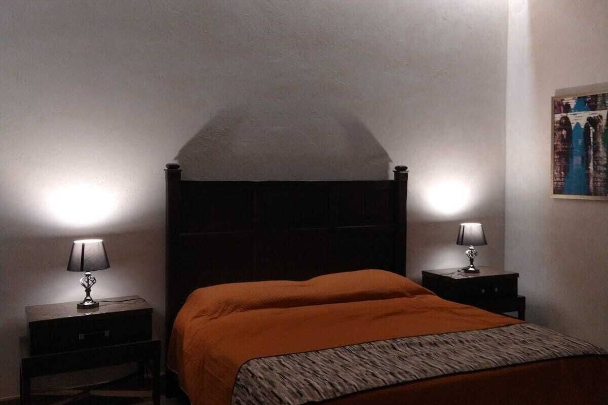 Une chambre avec un plafond en bois, un lit recouvert d’une courtepointe orange, deux tables de chevet avec des lampes et un petit tapis.
