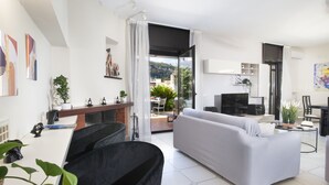 A modern living room with a grey sofa, a black armchair, and a dining area with a table and chairs. There is a kitchen area with a stove and cabinets, and a balcony with a view of the outdoors. - Maison Wally (Sorrento)