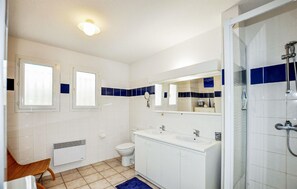 Une salle de bain avec des toilettes, un lavabo et une douche. Il y a deux fenêtres et un miroir au-dessus du lavabo.