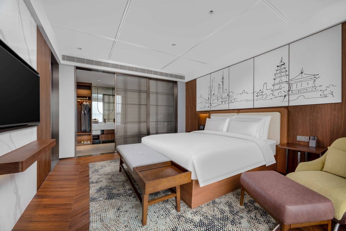 Een moderne hotelkamer met een groot bed, een flatscreen televisie, een bankje en een muur met een stadsgezichtsmural.
