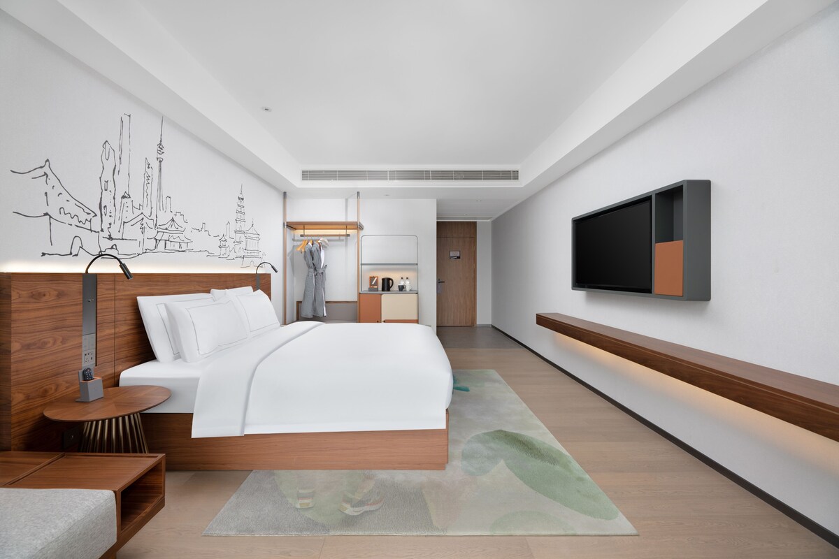 Een moderne hotelkamer met een groot bed, een aan de muur bevestigde televisie en een kleine kitchenette op de achtergrond.