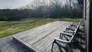 Una terraza de madera con una fila de sillas, un camino de ladrillo y una zona verde con árboles al fondo.