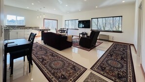 Un salón amplio con un sofá negro, una mesa de comedor con sillas, un televisor de pantalla plana y ventanas grandes con persianas.
