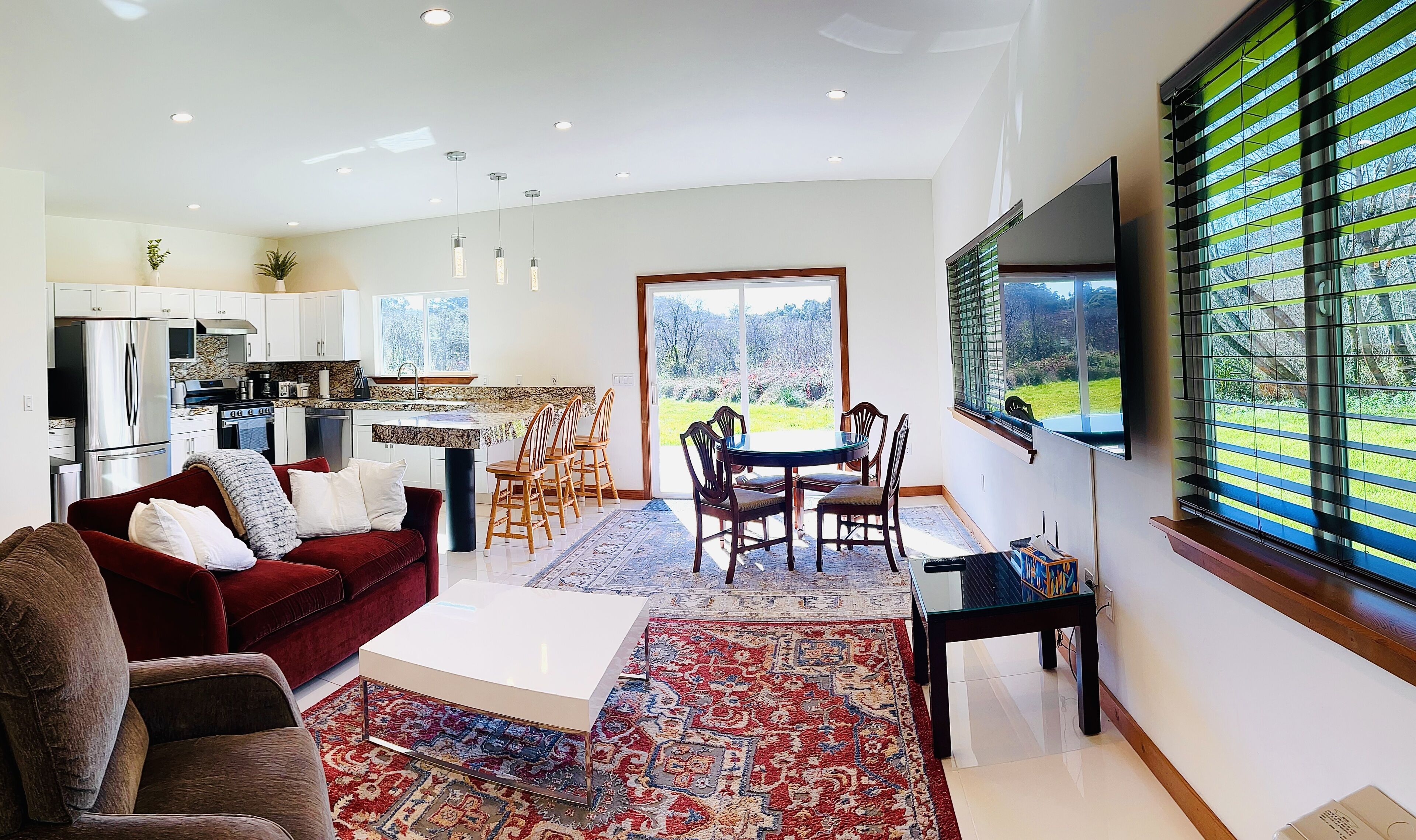 A modern living room with a red sofa, a coffee table, and a dining area with chairs and a table. A kitchen with white cabinets and appliances is visible in the background.