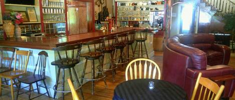 A bar area with wooden chairs and stools, a round table with a dark tablecloth, and shelves with various items.