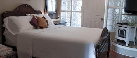 A neatly made bed with white linens and multiple pillows, a wooden headboard, a bedside table with a lamp, a mirror, and a television on a stand.