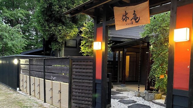 A traditional Japanese entrance with a wooden gate, stepping stones, and a hanging sign. - Kyuan Iida (Nasushiobara)