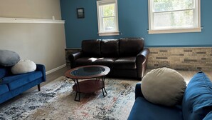 A living room with a blue wall, a brown leather sofa, a blue couch, a glass coffee table, and a ceiling fan.
