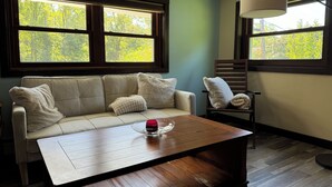 A cozy living room with a sofa, a wooden coffee table, and a chair. There are large windows with a view of trees.