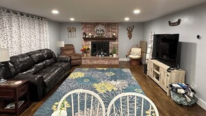 Ein Wohnzimmer mit einer Ledersofa, einem weißen Sessel, einem Fernseher, einem Kamin und einem gemusterten Teppich.