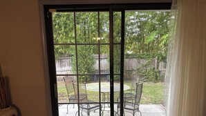 Vista a través de una ventana con celosía metálica, un patio con mesa y sillas, y una valla de madera al fondo.