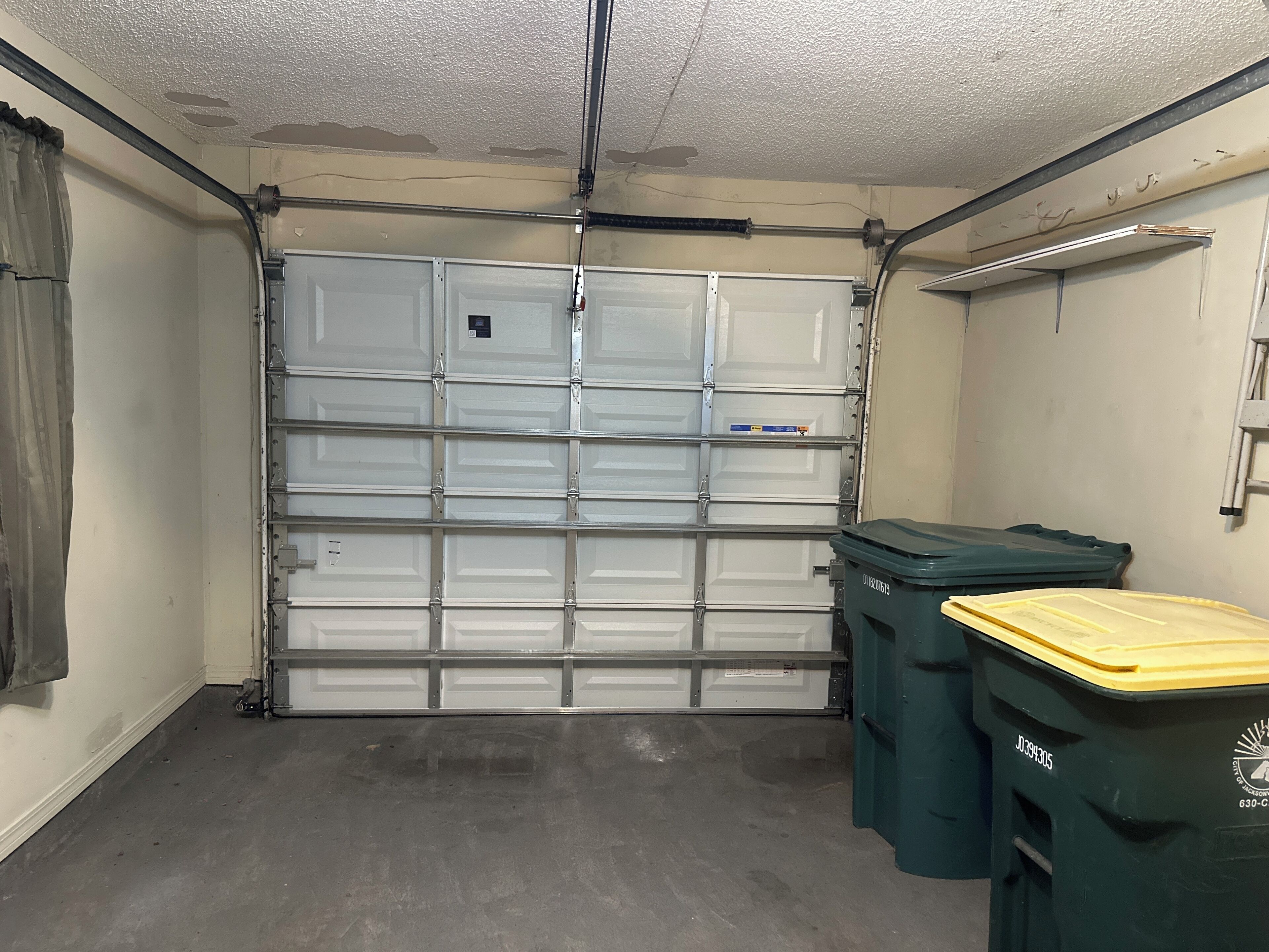 Eine Garage mit geschlossener Tür, einem Betonboden und zwei grünen Mülltonnen.