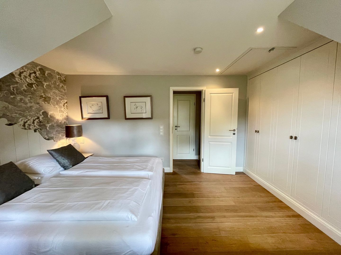 Quarto com duas camas, piso de madeira, guarda-roupa branco e porta que dá para outro cômodo.