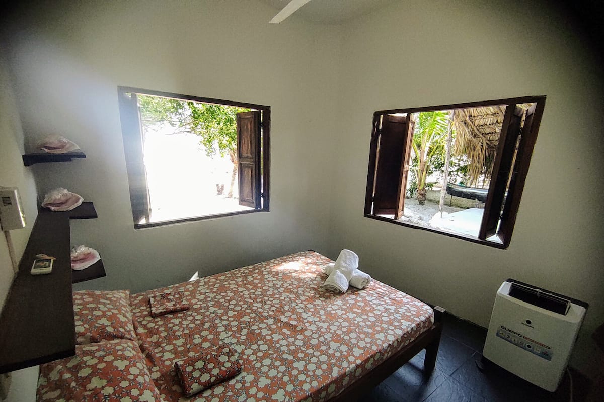 Quarto pequeno com uma cama de solteiro, cobertor estampado com flores, duas janelas com persianas abertas, aquecedor e um prateleira com produtos de higiene pessoal.