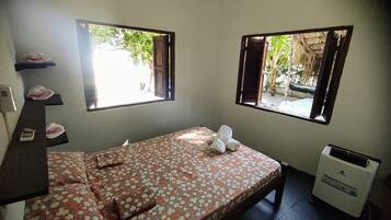Een kleine eenpersoonskamer met een bloemenprint bedspread, twee ramen met open luiken, een verwarming en een plank met toiletartikelen.