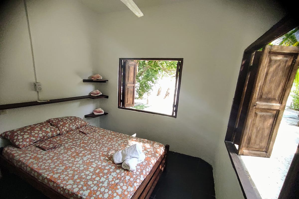 Quarto pequeno com cama de solteiro, janela, porta de madeira e prateleira com toalhas.