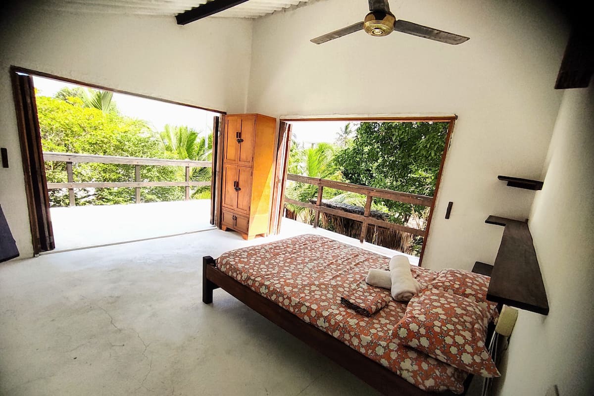 Quarto com cama, ventilador de teto e vista para o verde lá fora.