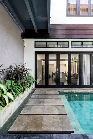 Une maison moderne dotée d’une piscine, d’un escalier en pierre menant à une porte en verre et d’un jardin où poussent différentes espèces de plantes.