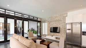 Een moderne woonkamer met een bank, stoelen, een salontafel, een aan een bakstenen muur bevestigde televisie en een koelkast.