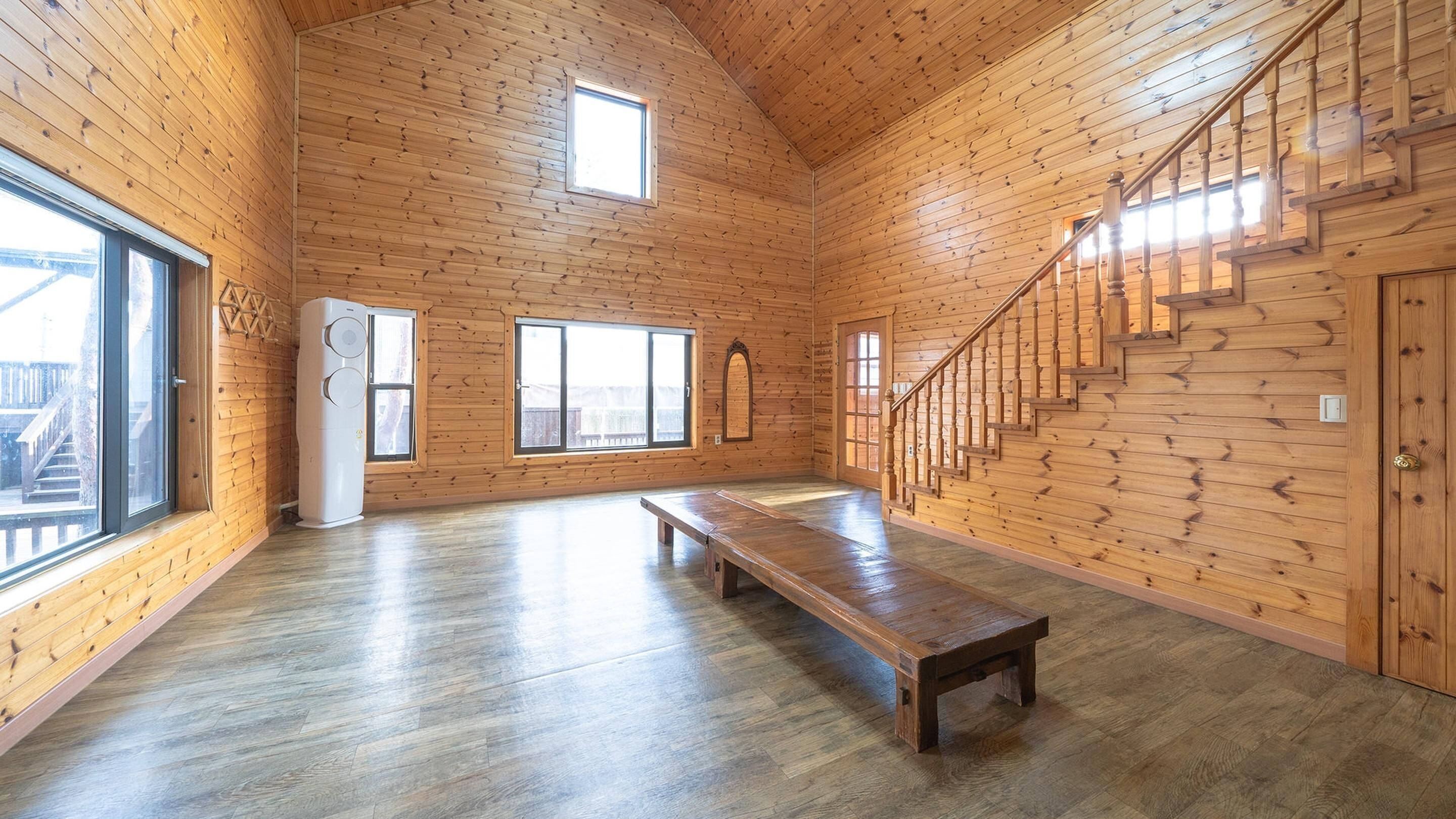 A spacious room with wooden walls and floors, a large window, a staircase, and a bench.