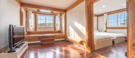 A room with a wooden floor, a bed, a desk, a television, and large windows.