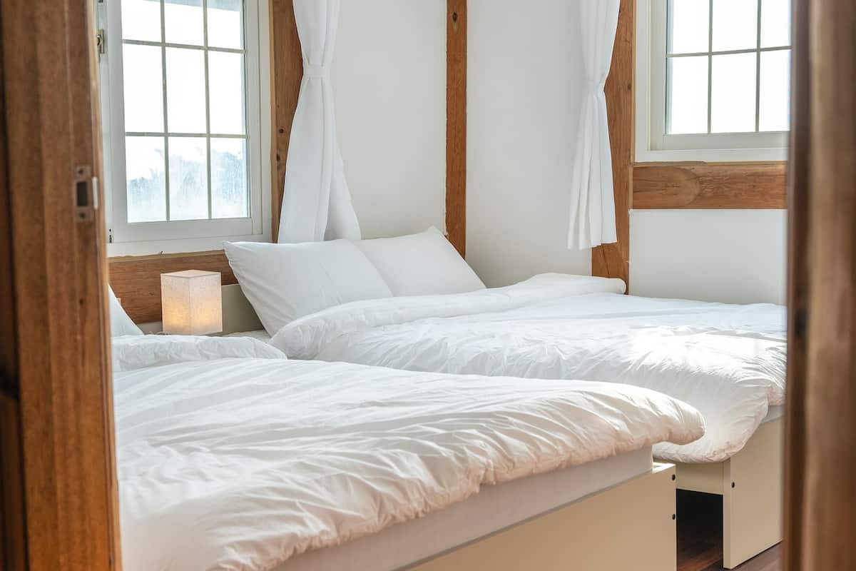 Une chambre avec deux lits, du linge de lit blanc, une table de chevet avec une lampe et des têtes de lit en bois.
