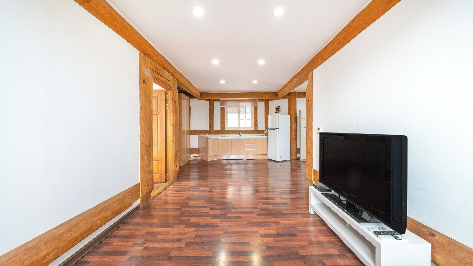 Corredor com piso de madeira que dá para uma cozinha equipada, com geladeira e televisão em móvel.