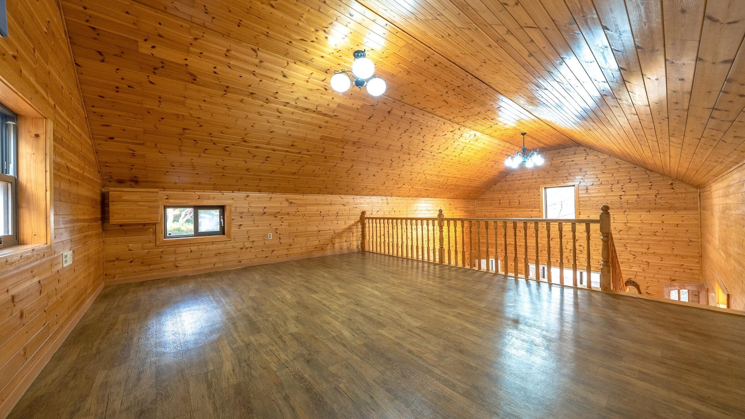 Um quarto amplo no sótão, com paredes e chão de madeira, com varanda, guarda-corpo e uma pequena janela.