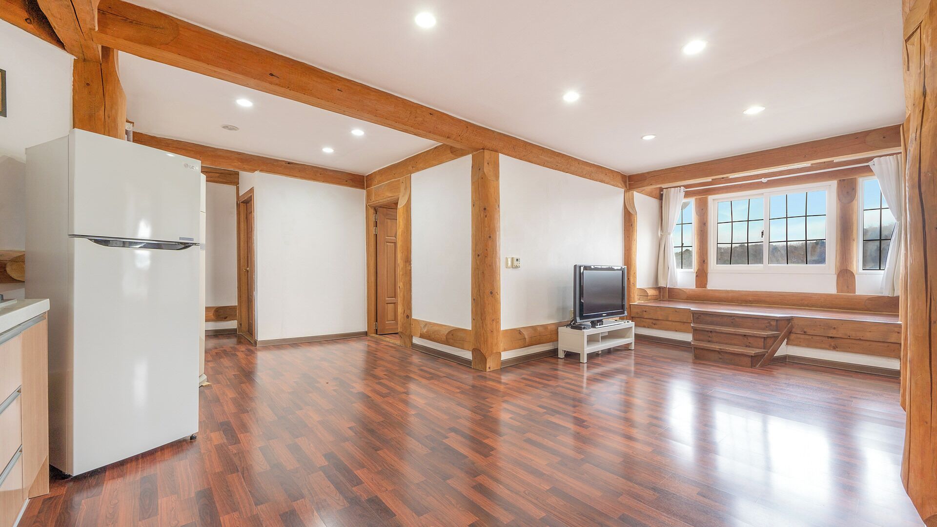 A spacious room with wooden beams, a white refrigerator, a television on a stand, and a window with a view.