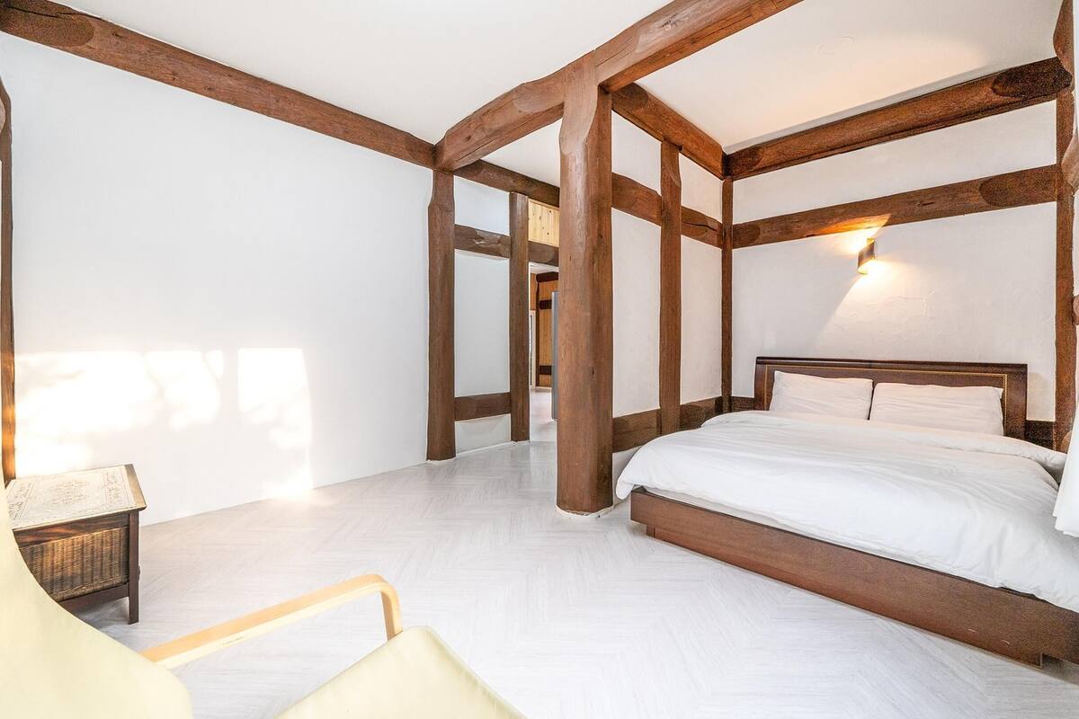 Une chambre avec des poutres apparentes, un lit recouvert de draps blancs, une table de chevet et une chaise.