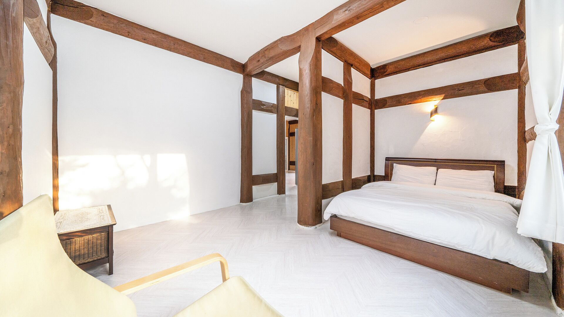 Quarto com estrutura de teto em madeira, cama com roupa de cama branca, mesa de cabeceira e uma cadeira.