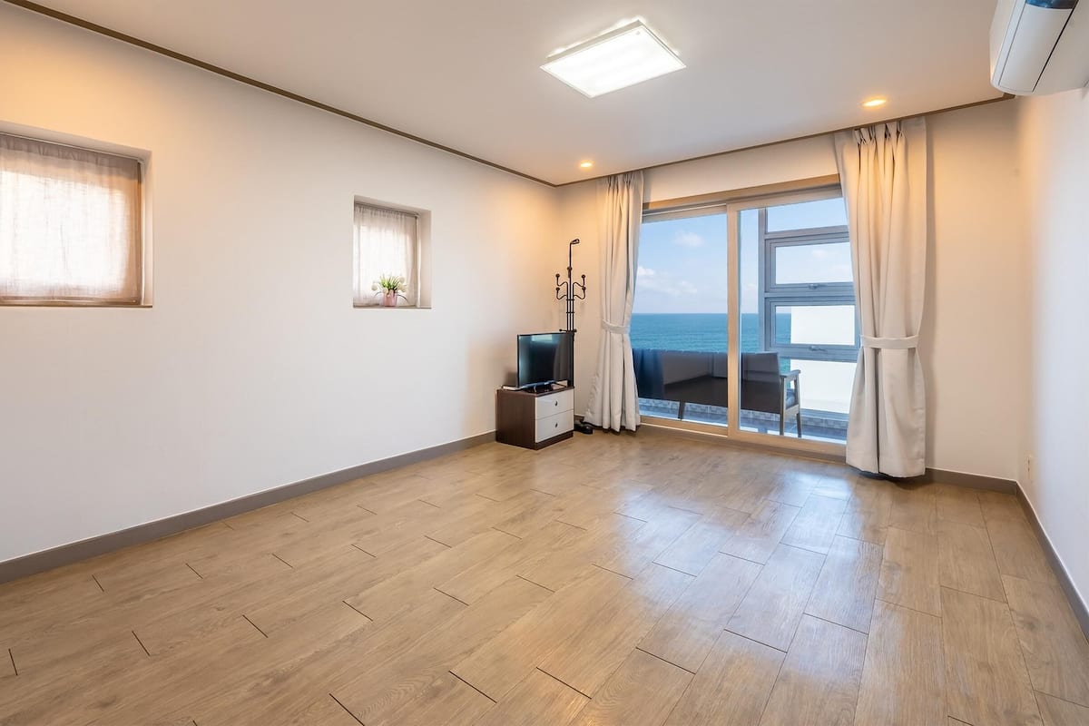 Una habitación amplia con suelo de madera, televisor, una mesita y ventana con vista al mar.