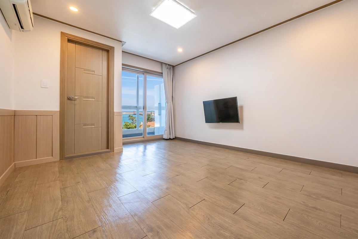Una habitación amplia con piso de madera, una puerta corredera de vidrio que da a un balcón con vistas, un televisor montado en la pared y un refrigerador blanco.