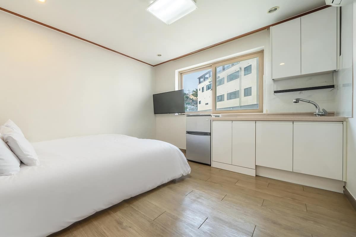 Una habitación compacta con cama, un pequeño kitchenette, televisor y ventana con vistas a edificios.