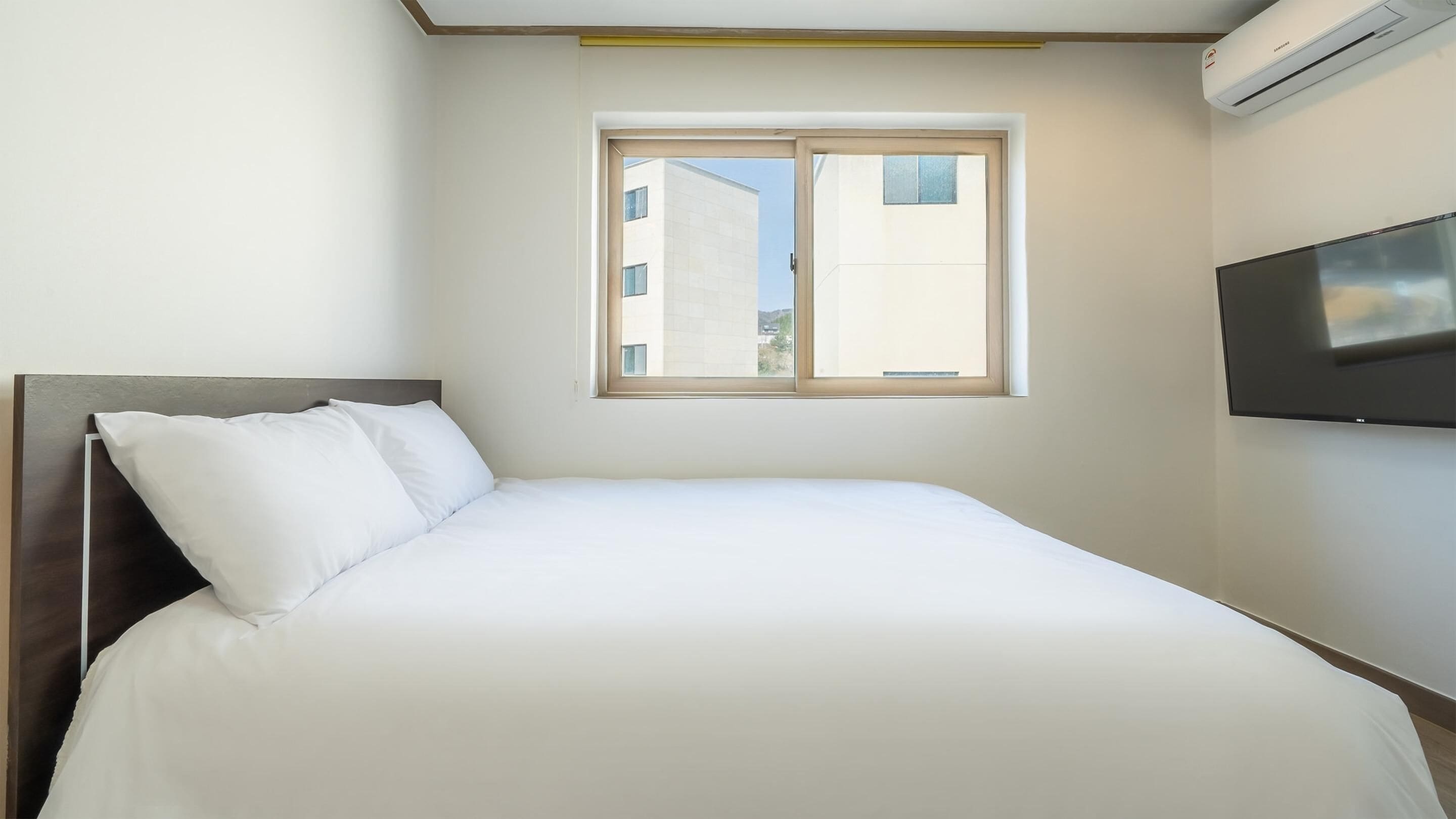 A neatly made bed in a room with a large window, a flat-screen TV mounted on the wall, and an air conditioning unit above the window.