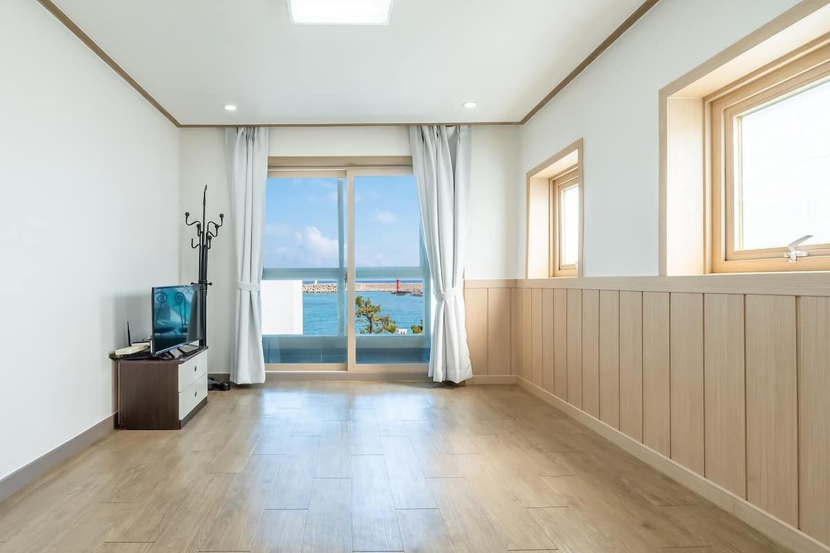 Una habitación amplia con suelo de madera, un ventanal con vistas al mar y un televisor sobre un mueble de madera.